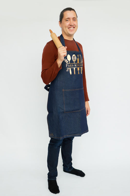 Mand med forklæde af mærk denim. På forklædet står der fars køkken i guld