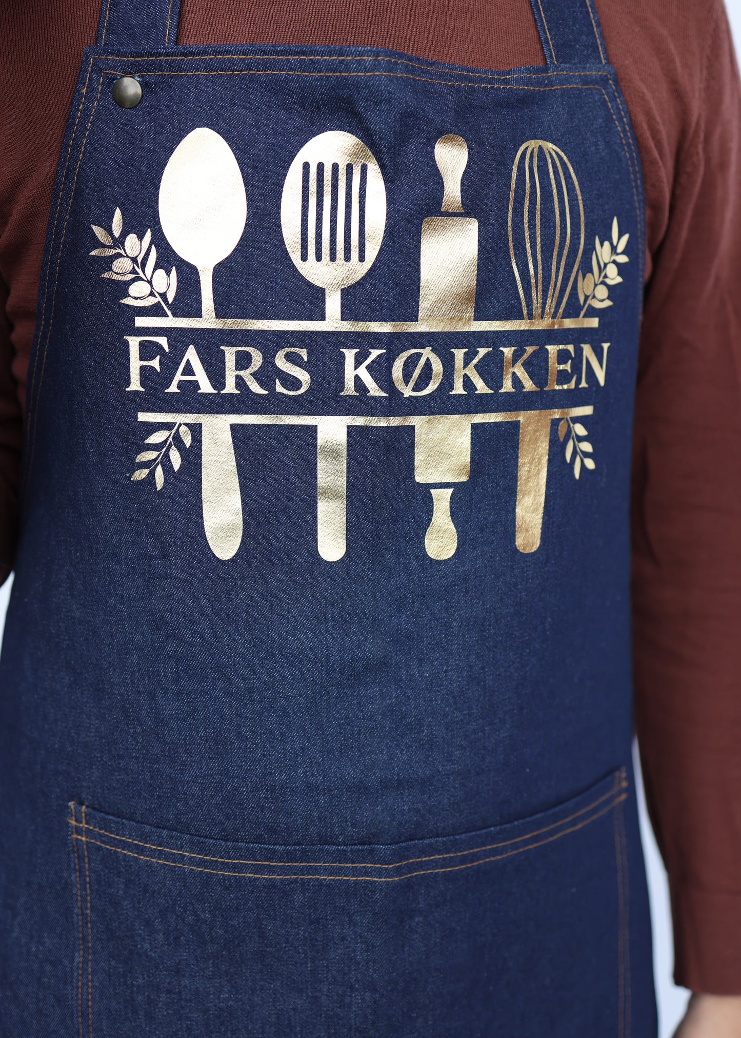 Zoomet helt tæt på denim forklædets tryk i guld, som forestiller en masse køkkenredskaber og navnet på manden
