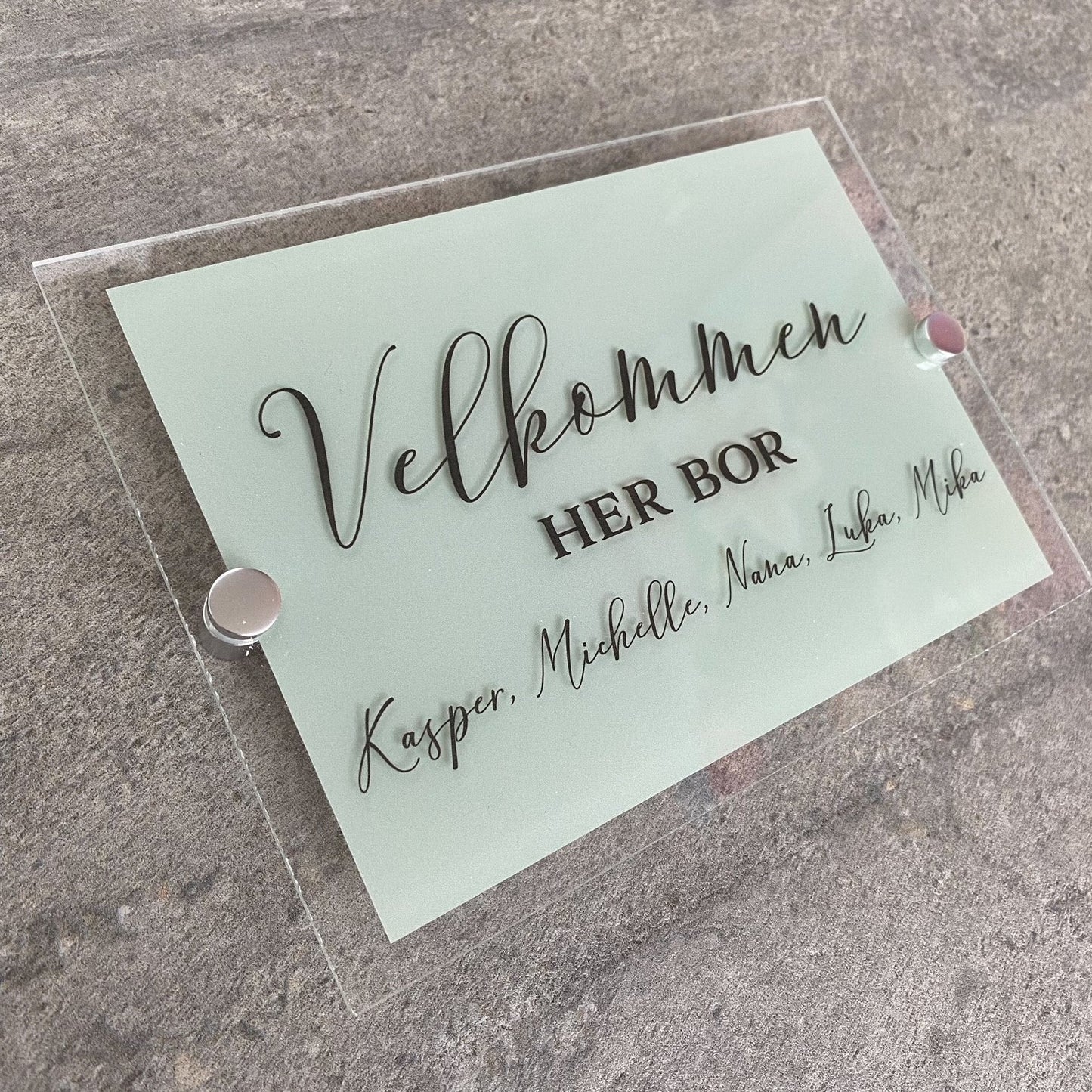 Dørskilt med navn. Lavet af akryl. Dette akryl dørskilt er lavet på klar akryl og har en stor firkant i lys grøn på bagsiden. Foran står der velkommen, her bor. Du skriver selv hvilke navne der skal stå på dørskiltet.