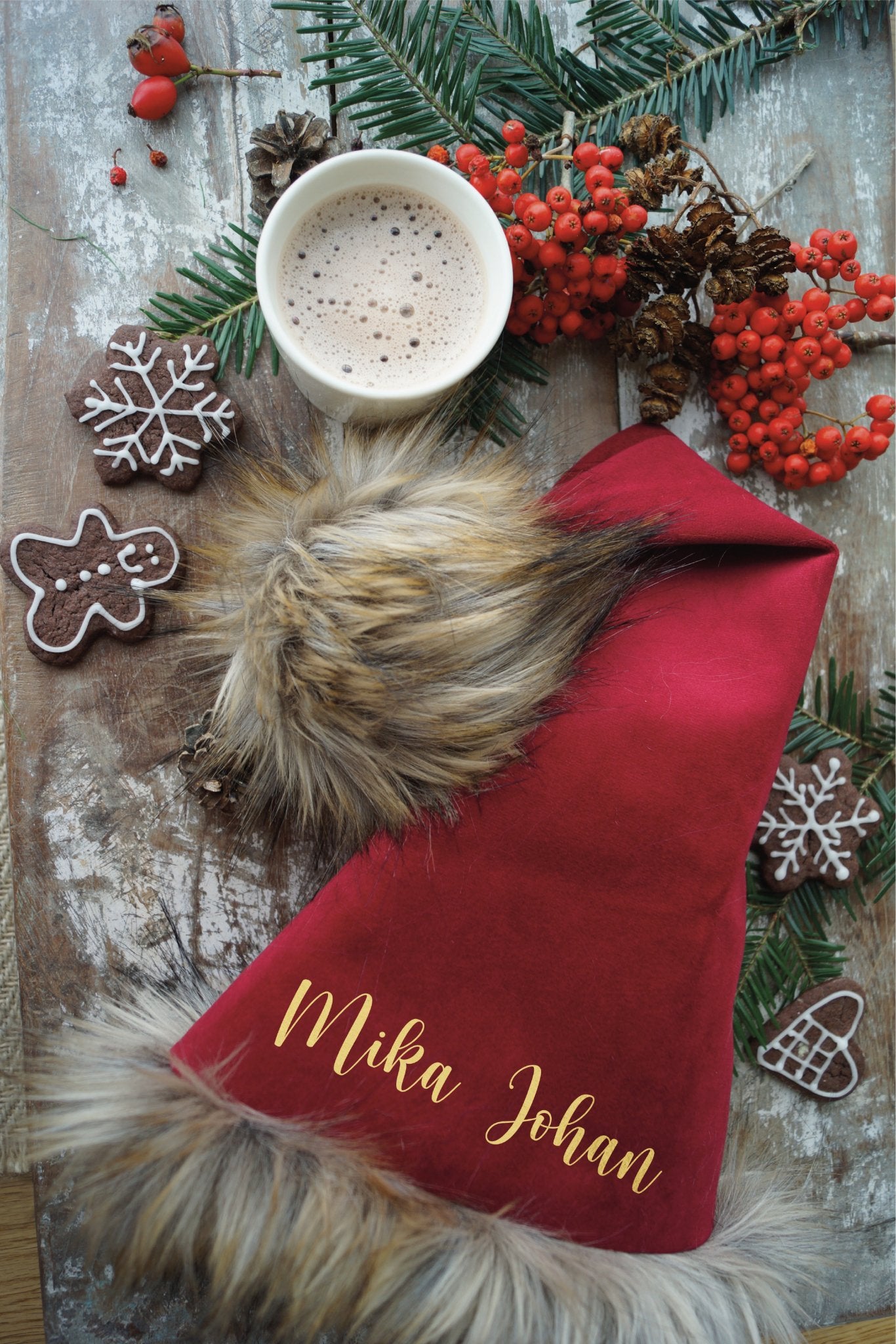 Smuk rød nissehue i høj kvalitet. Her ligger nissehuen på et julebord sammen med en kop varm kakao og små småkager. Nissehuen er lavet af velour og har en pelskant og kvast i imiteret ulvepels. Navnet er lavet i guldfolie